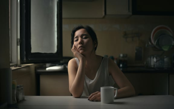 A woman sits thoughtfully at a kitchen table with a mug.