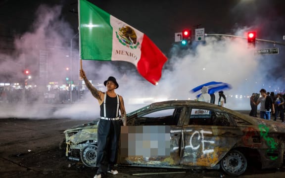 EDITORS NOTE: Graphic content / A protester waves the Mexican flag beside the wreckage of a burnt car sprayed with graffiti against US Immigration and Customs Enforcement (ICE), as law enforcement clashes with demonstrators during a protest following federal immigration operations, in the Compton neighborhood of Los Angeles, California on June 7, 2025. US President Donald Trump deployed 2,000 troops on June 7, 2025 to handle escalating protests against immigration enforcement raids in the Los Angeles area, a move the state's governor termed "purposefully inflammatory." Federal agents clashed with angry crowds in a Los Angeles suburb as protests stretched into a second night Saturday, shooting flash-bang grenades and shutting part of a freeway amid raids on undocumented migrants, reports said. (Photo by RINGO CHIU / AFP)
