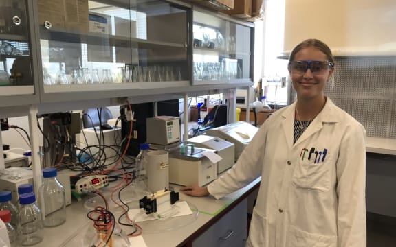 Liz stands in a lab with a white lab coat and lab goggles on. She has her hand on the bench beside some equipment, including a centrifuge and DNA gel electrophoresis set up.