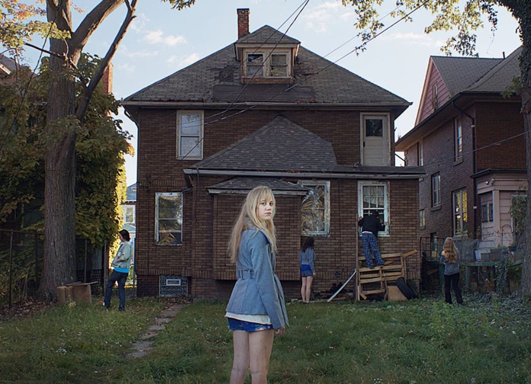 A young woman stands in front of a shadowy and dilapidated brick house.