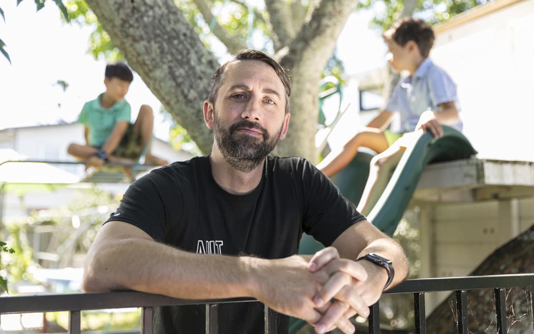 A portrait photo of Scott from torso up. He is smiling at the camera wearing a black t-shirt. His arms are draped over a metal fence, fingers interlaced. Behind him is the out-of-focus background is a playground, with a large tree and two children playing on a slide and a climbing frame.