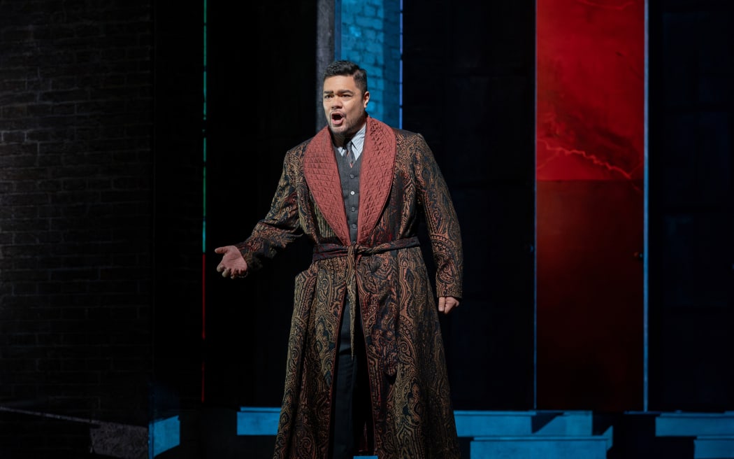 Pene stands on stage in a rich paisley robe draped over a suit. He is singing and gesturing.