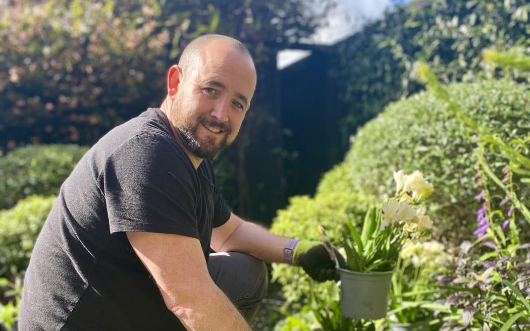 Martin is wearing a black t-shirt. He is kneeling down by a flower bed, holding a pot with a plant in. He is looking towards the camera and smiling.