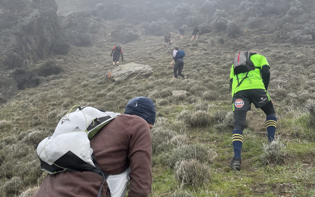 People in outdoors gear and wearing backpacks climb up a steep tussocked mountain side towards the foggy peaks,