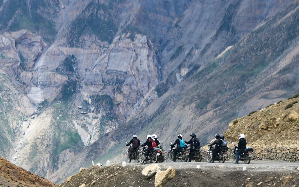 A group of motorcycle riders are on a small road halfway up a mountain. The mountain towers behind them.