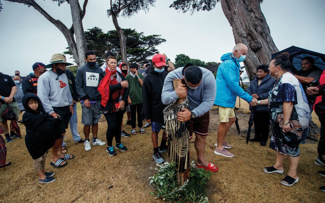 A man embraces a wooden post draped with a cloak and with wreaths of vegetation at its base. The grass is dry and brown, and people wearing caps and some with face masks gather around in a rough circle. A woman shakes the hand of a man to the right.