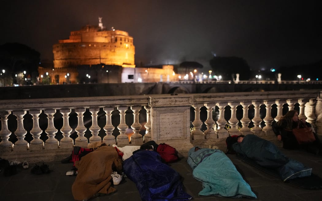 People rest in sleeping bags on the street ahead of late Pope Francis' funeral with a view of the Mausoleum of Hadrian near the Vatican, in Rome on April 26, 2025. Pope Francis, champion of the poor and the Church's first Latin American leader, will be honoured April 26 with a funeral attended by world leaders and tens of thousands of Catholic faithful. The Argentine pontiff, who died on April 21, 2025, aged 88, sought to create a more open-minded Church during his 12-year papacy, and many emotional tributes have been made -- with 250,000 people paying respects at his coffin in St Peter's Basilica. (Photo by Dimitar DILKOFF / AFP)