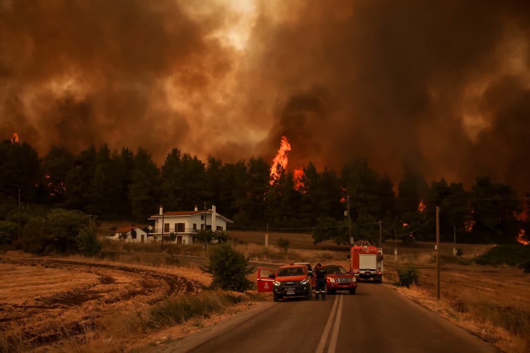 Firefighters stand on a road as flames rise from a forest in the background in the village of Kyrynthos, in the north of Evia Island .