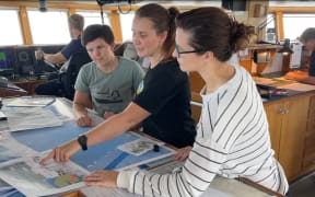 Three women look at a map laid out on a table in a room inside a ship. The middle woman is pointing at something. Two people are engaged in other tasks in the background.