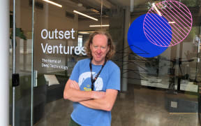 Sean smiles at the camera. He is standing outside an office building. The window of the business says "OUTSET VENTURES - THE HOME OF DEEP TECHNOLOGY".