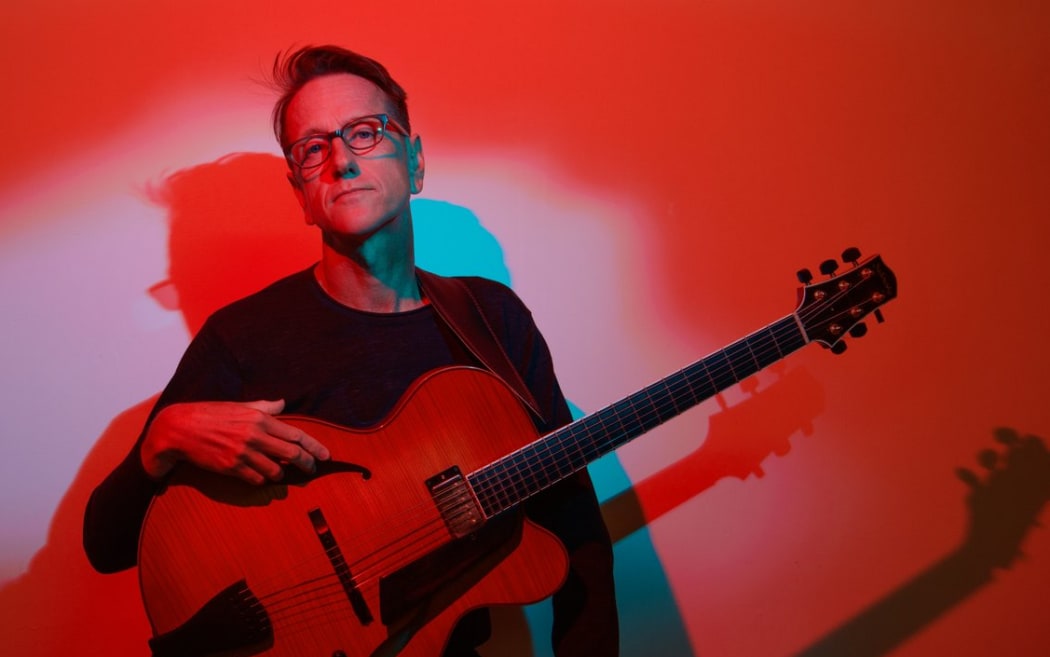 A red-toned photo of guitarist James Sherlock, holding an archtop guitar and looking at the camera