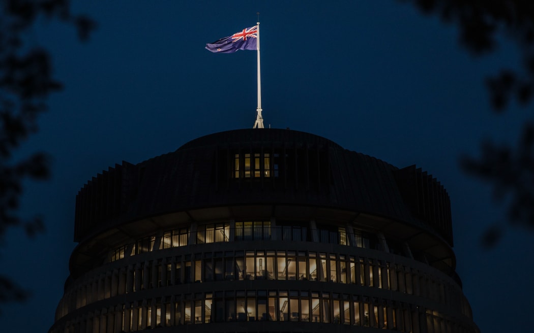 A night view of the Beehive, with interior lights on. The New Zealand flag is flying atop the building against a dark blue sky, framed by silhouetted tree branches.