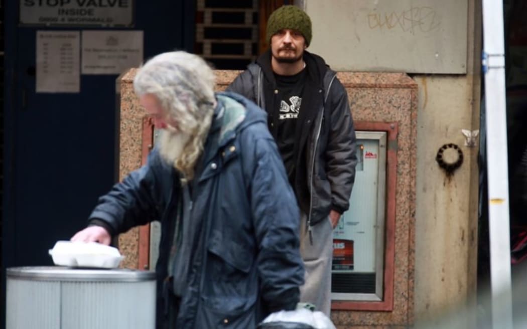 As Jason  begs, an old man in foreground looks though rubbish bin