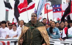 Ngātiwai leader Aperahama Edwards walks solemnly at the front of the hīkoi mō te Tiriti group across Auckland's Harbour Bridge He wear a traditional pākē harakeke (Māori rain cape), used for both protection and shelter.