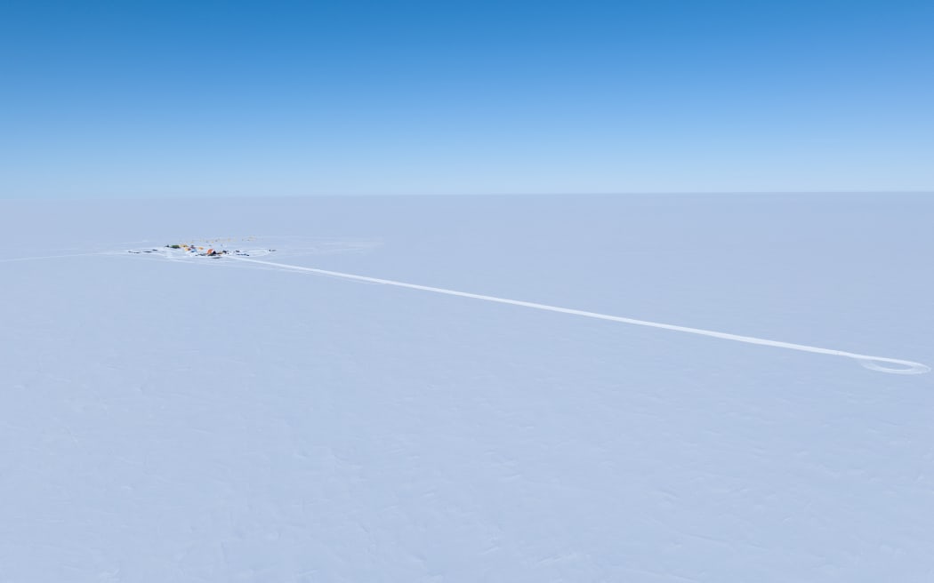 A drone image of the white expanse of flat ice beneath a clear blue sky with a tiny camp off to the left, and a long trail in the ice extending to the right.