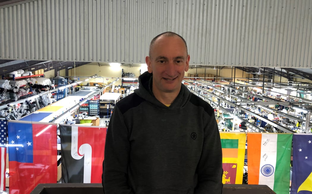 Ben Kepes wears a black hoodie and stands on an elevated platform. Behind him is the factory floor of Albion Clothing, with rows of workbenches and tools. There is a string of flags hanging over the factory floor.