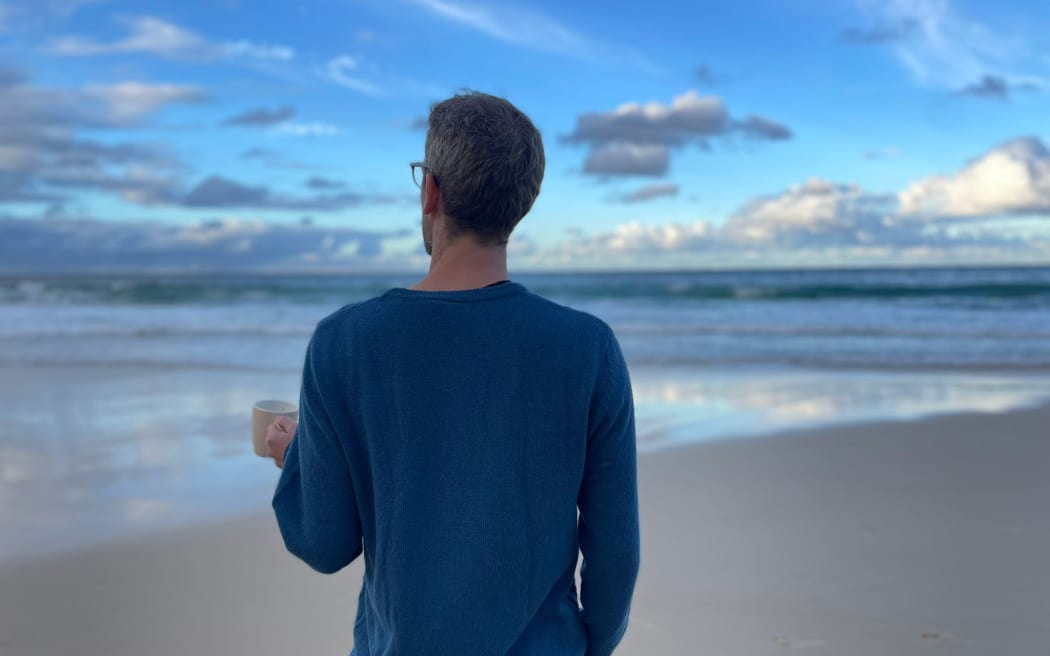 A man wearing a blue long-sleeved t-shirt stand on a beach holds a mug in his left hand while staring out to sea. The sky is mostly blue filled with light cloud.