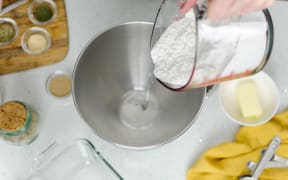 A person pouring flour into a bowl. On the table is a small bowl of butter, measuring spoons and small pots of spices.