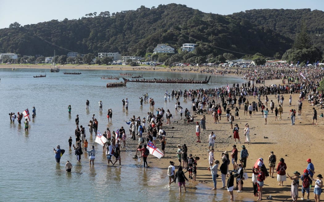 Hundreds of people standing on the beach, some wading into the water to greet the kaihoe in the Waka parade.