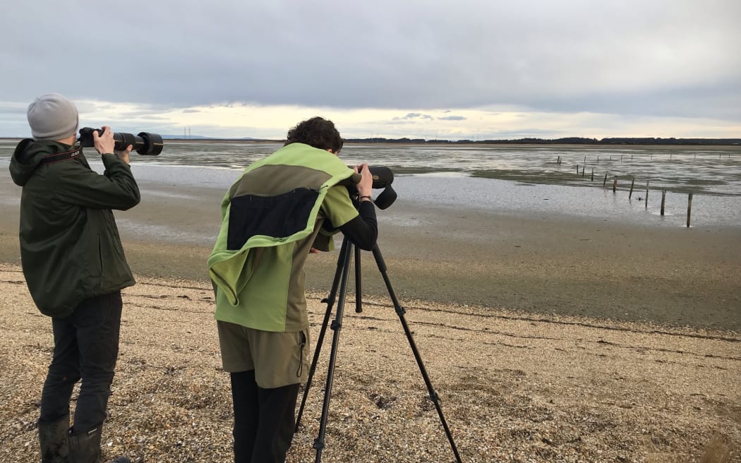 Two people wearing warm clothes are standing with their backs to the camera, looking out into an expanse of mudflat and shallow estuary. One person is wearing a beanie and has a long lens camera. The other person is wearing green and peering through a scope on a tripod. It is a grey overcast sky.