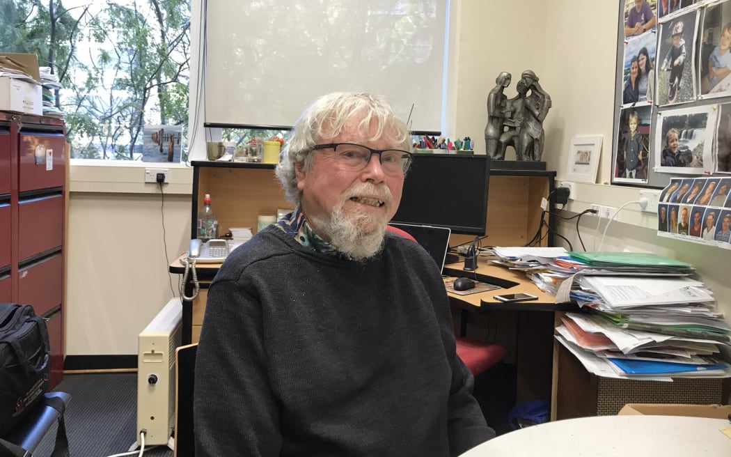 Warren Tate sitting at a desk in his office with photos on the wall and files on the top of shelves and tables behind him.