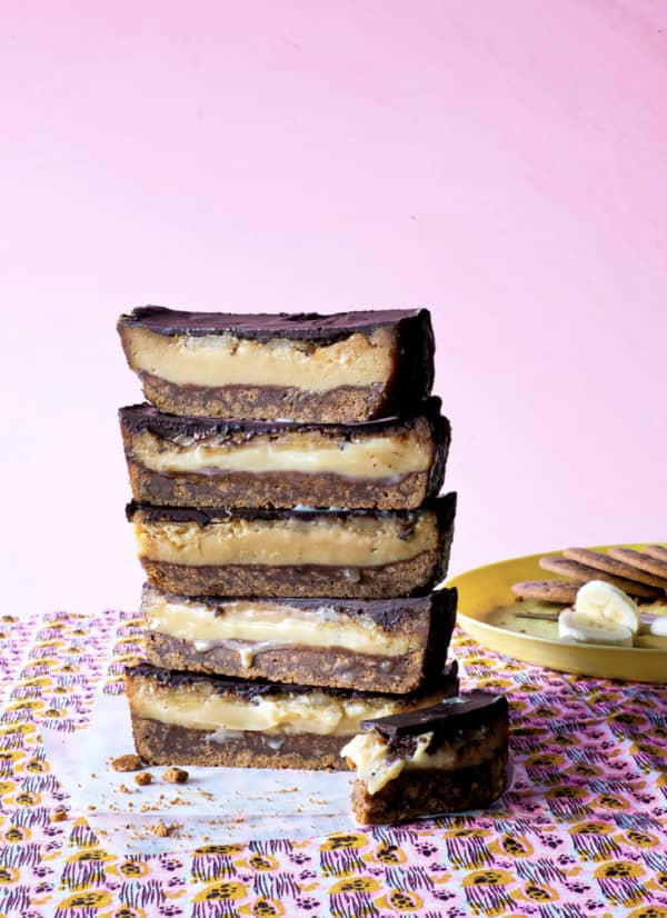 A vertical stack of five pieces of banana caramel slice, showing a cross-section of biscuit base, gooey caramel and chocolate topping, on a pink patterned cloth.