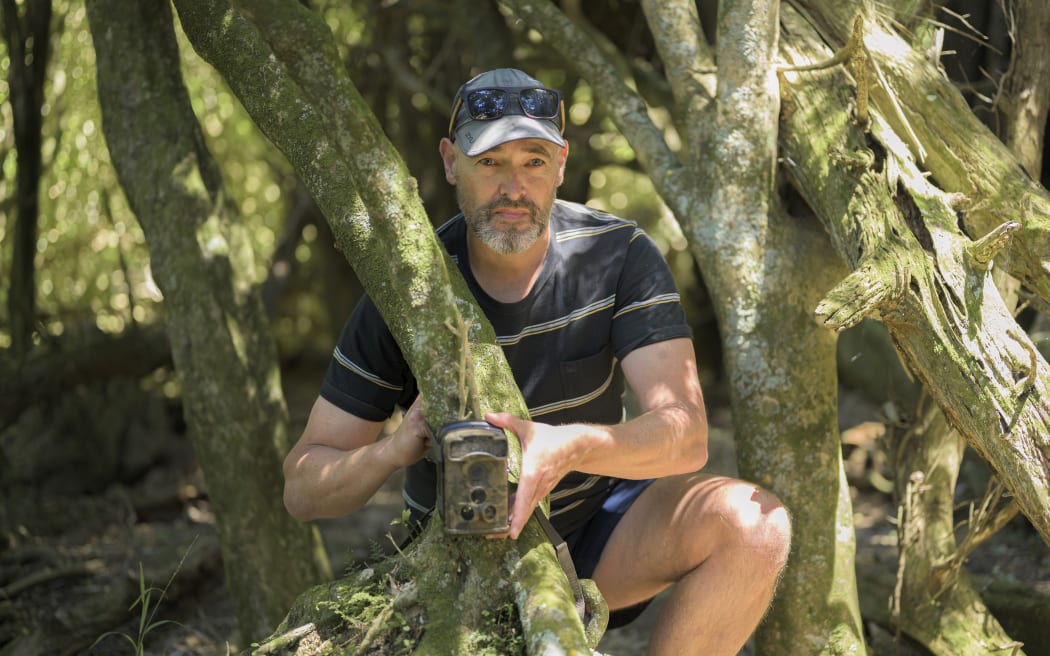 A man wearing a cap stands behind a tree trunk with a camera trap attached to the trunk.