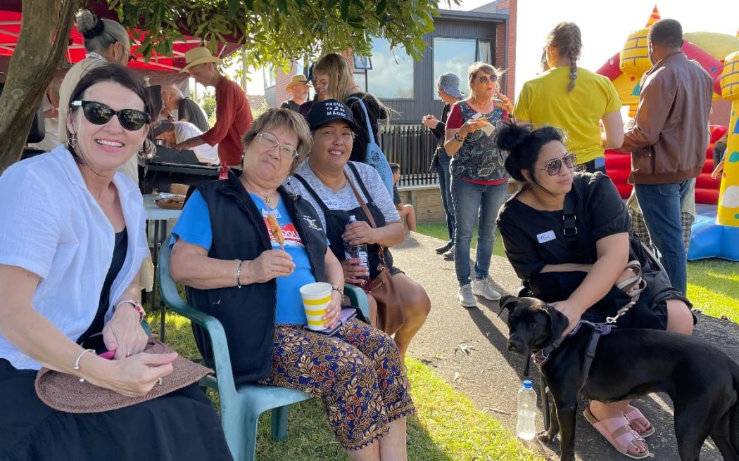A group of people sitting on green plastic deck chairs carrying plastic cups and smiling at the camera. There are many more people barbecuing and chatting in the background.
