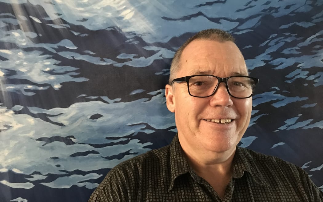 A man standing in front of a watery background. He is wearing glasses and smiling.