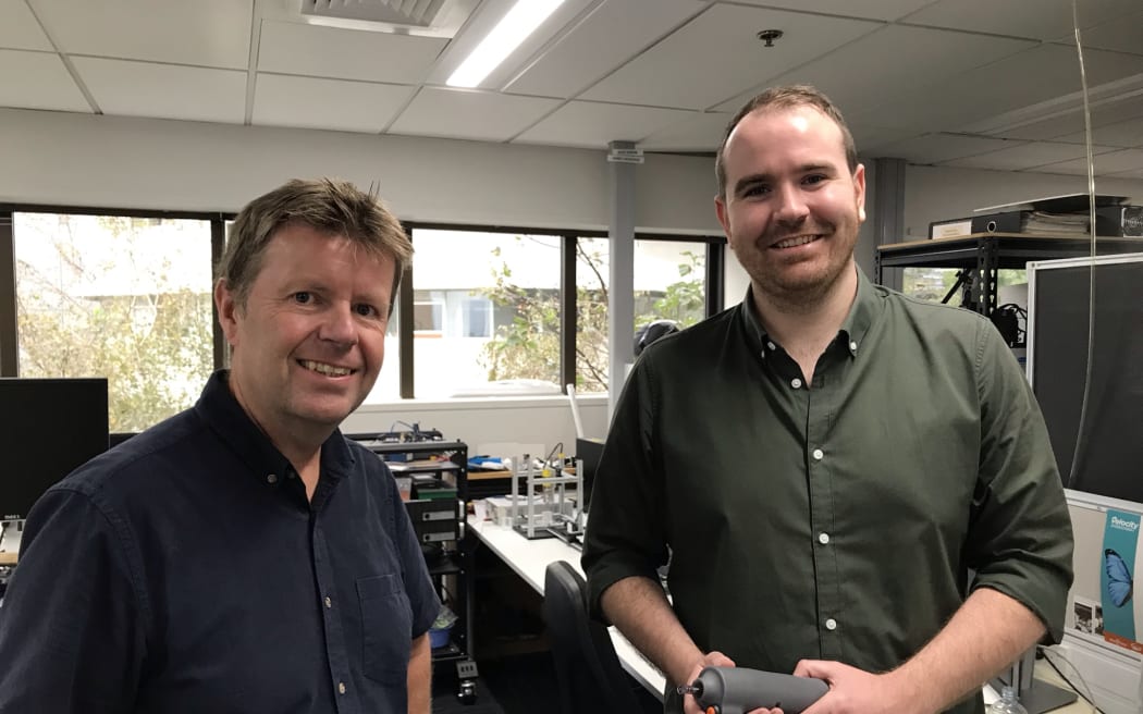 Andrew and James stand next to each other, James has the research version of the electric motor jet-injector in his hand - its a cylindrical grey shape.