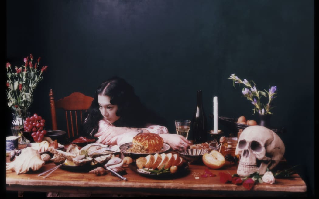 Singer and author Michelle Zauner poses leaning on a wooden table which is spread with a skull, candle and various food items.