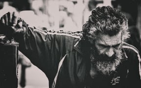 It is a black an white photo of a man (possibly homeless, but very stylish) leaning on a wall looking down towards the ground.