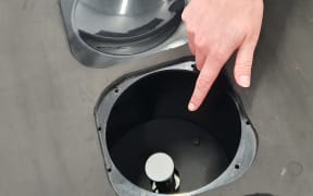 A close-up of a hand pointing to a circular metal hole in a surface, with a white disk inside the hole.