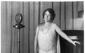 New Zealand-born opera singer Frances Alda, full length, standing, between piano and radio microphones, facing left.
