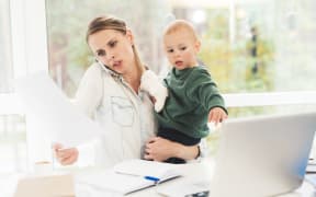 A photo of a woman who is working from home while she cares for a child at the same time. A woman with a child is sitting in a bright room.
