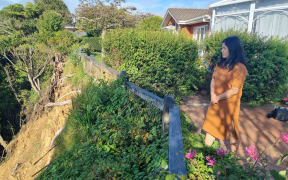 Jennifer Chen has black long hair and wears an orange dress. She stands at the edge of her property looking down at the cliff which has fallen away.
