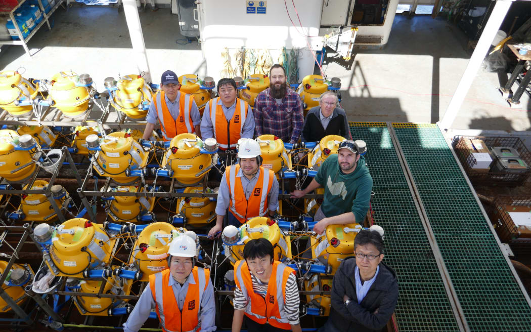A group of nine people wearing hard hats and hi-vis vests gather next to large yellow cylindrical devices in metal frames on the deck of a ship.