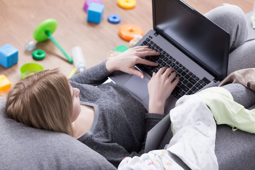 Young and tired woman lying on sofa with laptop, toys lying on the floor