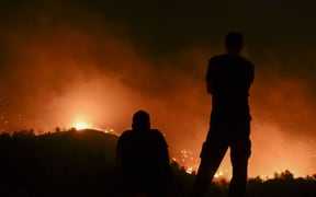 People watch the fires near the village of Malona in the Greek island of Rhodes on July 23, 2023. Tens of thousands of people fled wildfires on the Greek island of Rhodes on July 23, 2023, as terrified tourists scrambled to get home. Firefighters tackled blazes that erupted in peak tourism season, sparking the country's largest-ever wildfire evacuation -- and leaving flights and holidays cancelled. (Photo by SPYROS BAKALIS / AFP)