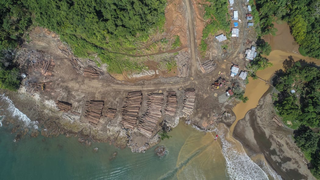 Aerial photo of the coast of the Solomon Islands showing a log landing area with lots of piles of logs waiting to be picked up by boat, plus some logging machinery and huts
