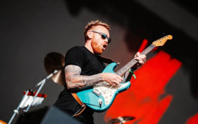 Joel Shadbolt, a red-headed and bearded man in a black t-shirt, raises his teal blue electric guitar to the sky.