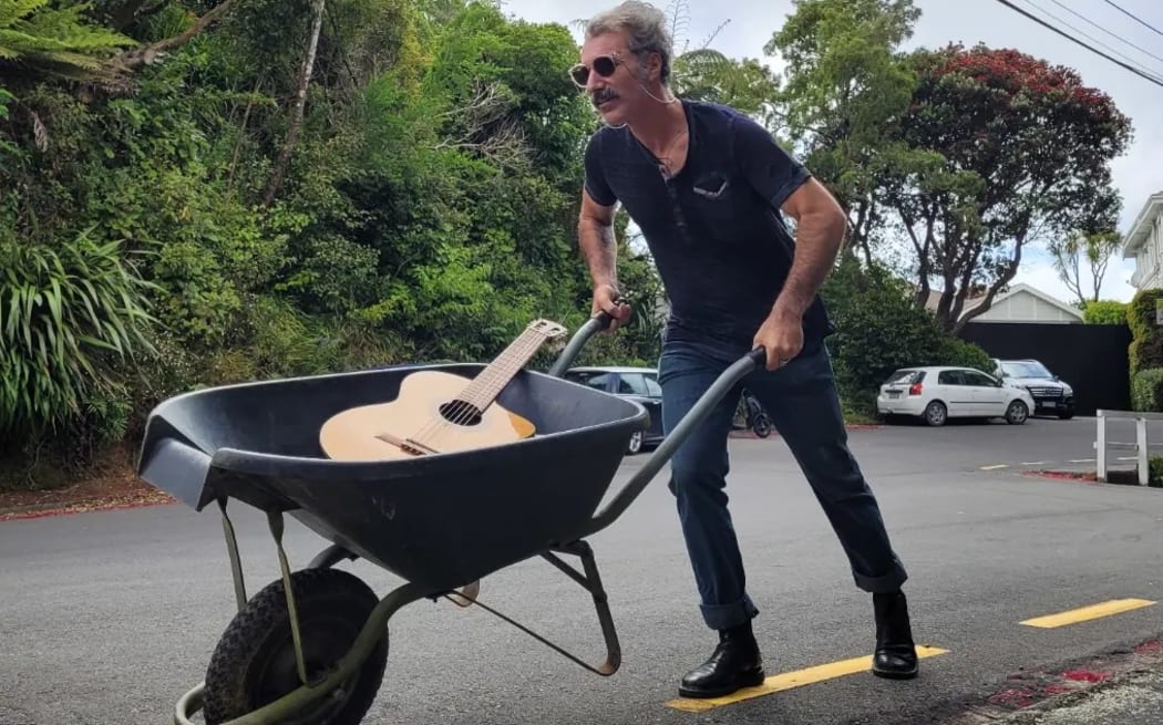 Samuel wears black sunglasses and carries a wheelbarrow with an acoustic guitar down a residential road.