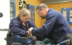 Richard assists a student with a piece of machinery. They both wear navy worksuits.