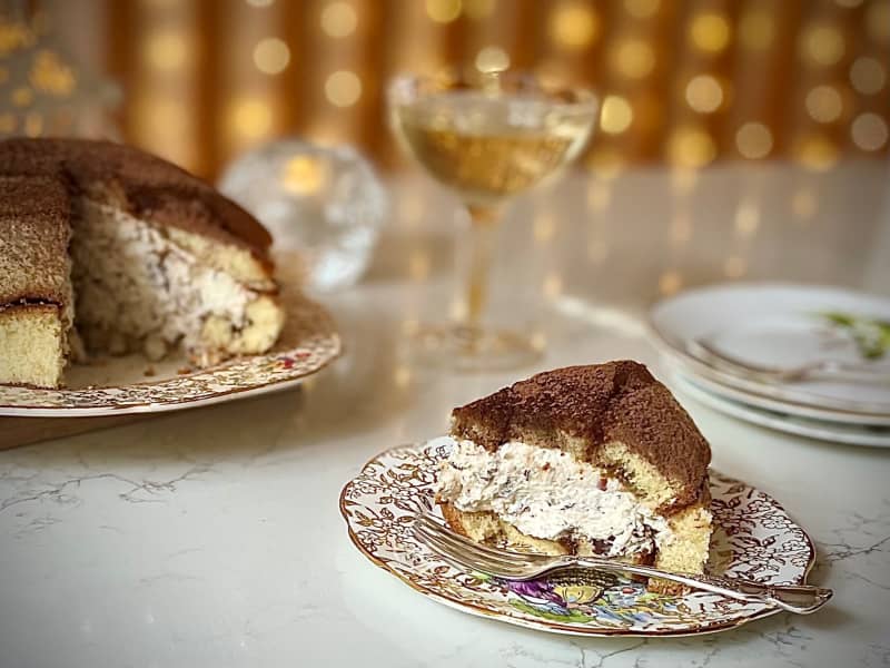 A whole Zuccotto (a dome of sponge filled with whipped cream laced with chocolate and hazelnuts) sits on a plate with a wedge cut out of it, the wedge is on a plate in the right foreground. There is a glass of prosecco and fairy lights in the background.