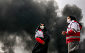 Women members of Iran's Red Crescent society stand near smoke plumes from an ongoing fire following an overnight airstrike on the Shahran oil refinery in northwestern Tehran on March 8, 2026. The United States and Israel launched strikes against Iran on February 28, sparking swift retaliation by the Islamic republic which responded with missile attacks across the region. The war has dragged in global powers, upended the world's energy and transport sectors, and brought chaos to even usually peaceful areas of the volatile region. (Photo by AFP)
