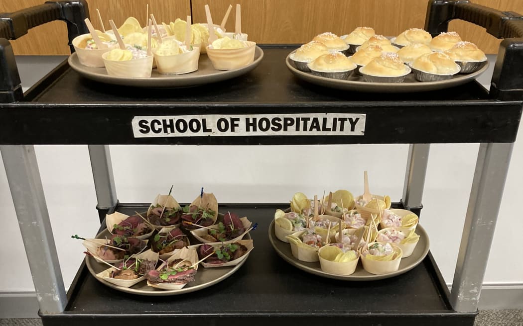 A trolley labelled School of Hospitality which is carrying six platters of food; kokoda, venison, duck and coconut buns.