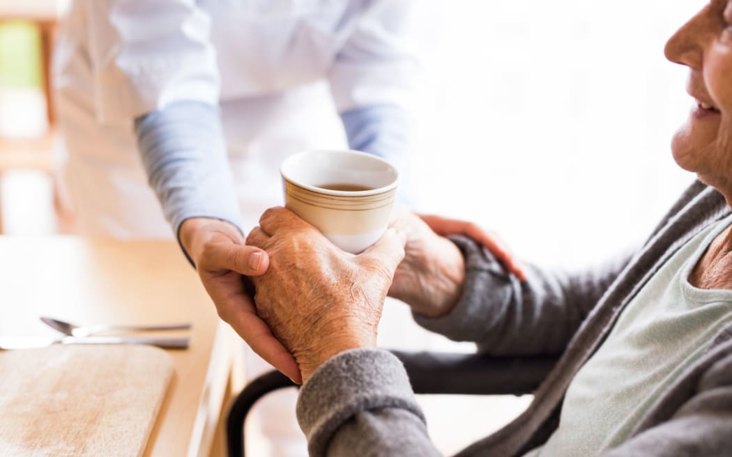 Unrecognizable health visitor and a senior woman during home visit. A nurse giving tea to an elderly woman sitting at the table. Close up.
