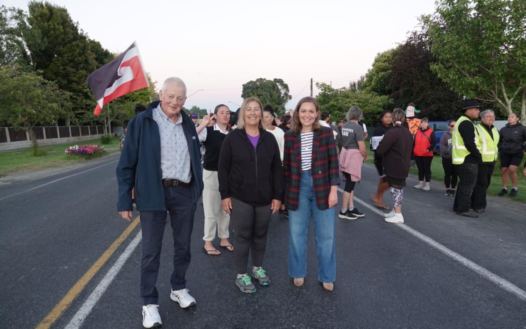 Wairoa mayor Craig Little, deputy mayor Denise Eaglesome-Karekare and local MP Katie Nimon stand in front of a group of people on the road. A tino rangatiratanga flag is flying behind them.
