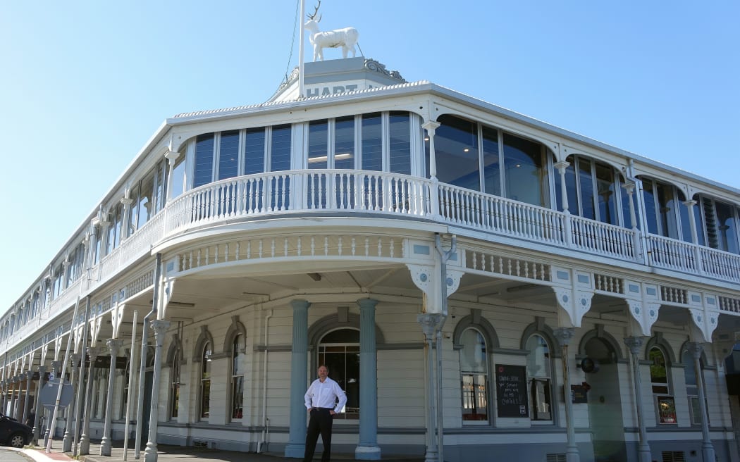 Man stands  outside front door on covered veranda of restored  2 story  early twentieth century pub.