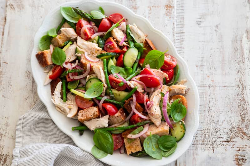 An oval serving dish containing a salad of toasted bread, basil, asparagus, red onion, tomatoes and rotisserie chicken.